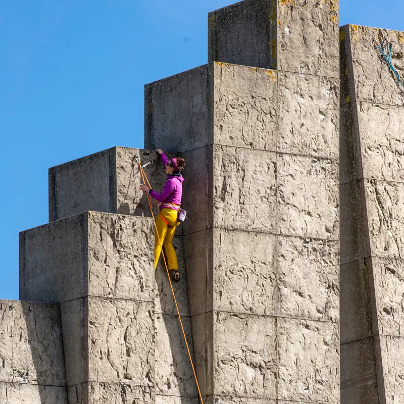 Tegen een helderblauwe lucht klimt iemand in een paars jasje en een gele broek met behulp van een touw tegen een groot betonnen bouwwerk op.